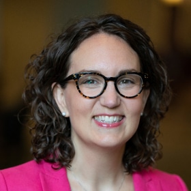 A woman with curly brown hair and dark glasses. She has on a hot pink jacket.